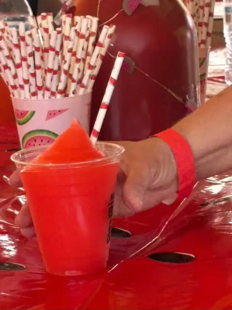 A person holding a bright red, watermelon slushie in a plastic cup at a sunny outdoor fair.