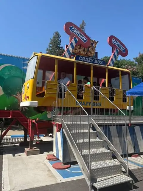 Children riding a "Crazy Bus" mechanical amusement park ride spinning in the air under a blue sky.