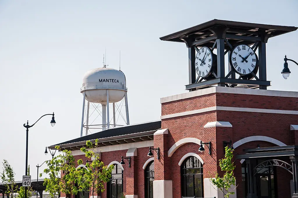 The brick building of the Manteca Transit Center featuring a prominent black clock tower, with the white Manteca water tower standing visible in the background against a clear blue sky.